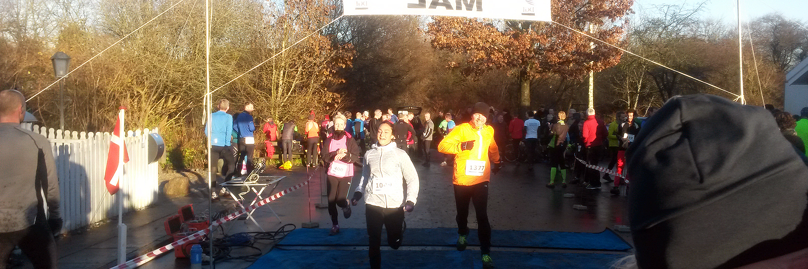 3 løbere i lange bukser løber om kap under mål-banner i skarp sol med skov i baggrunden